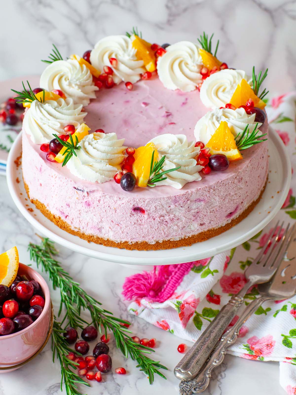 A pink cranberry cheesecake garnished with cream and fruit, on a cake, on a table with fresh rosemary.