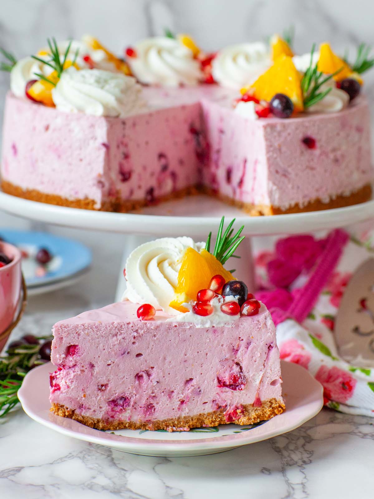 A slice of cranberry cheesecake on a plate, with a cheesecake on a cake stand in the background.