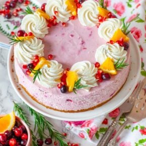 A cranberry cheesecake on a cake stand - topped with whipped cream and fruit.