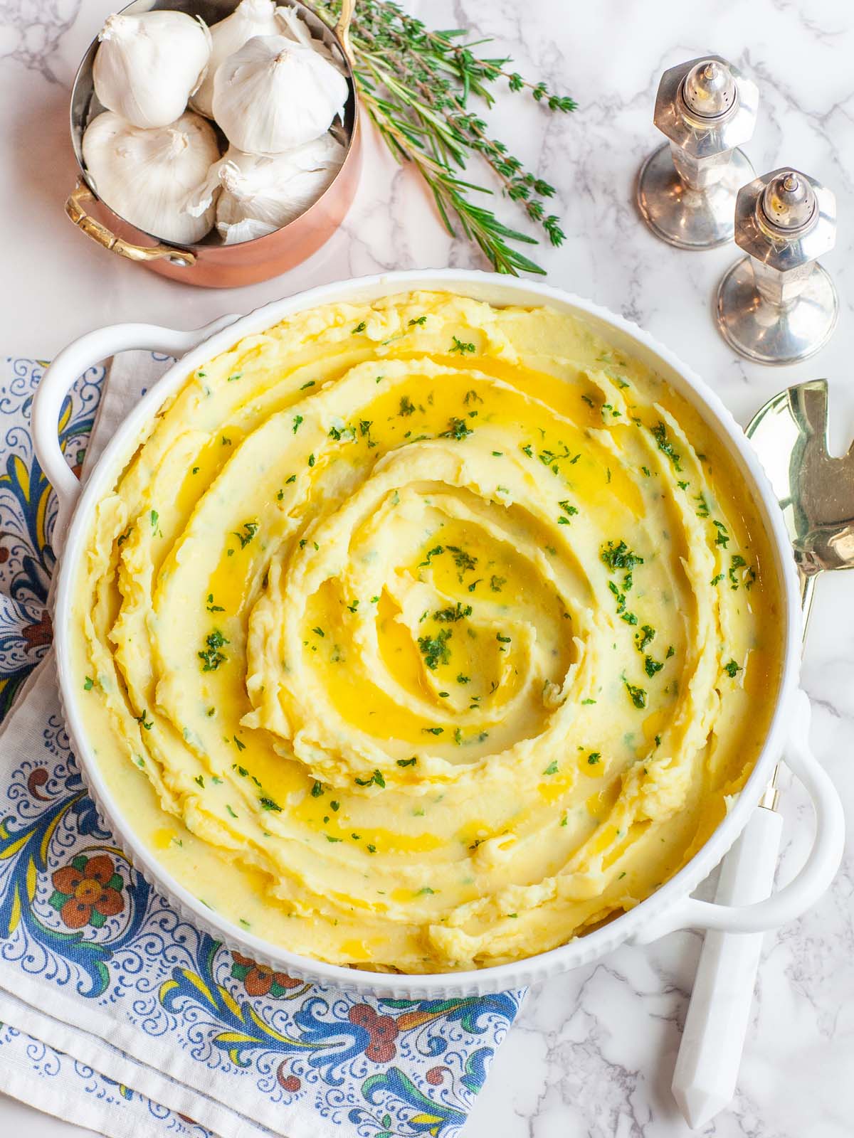 A large serving bowl of creamy buttered mashed potatoes with herbs, on a table with garlic and salt and pepper shakers.