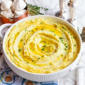 A closeup image of mashed potatoes in a white serving bowl, on a table with kitchen utensils.