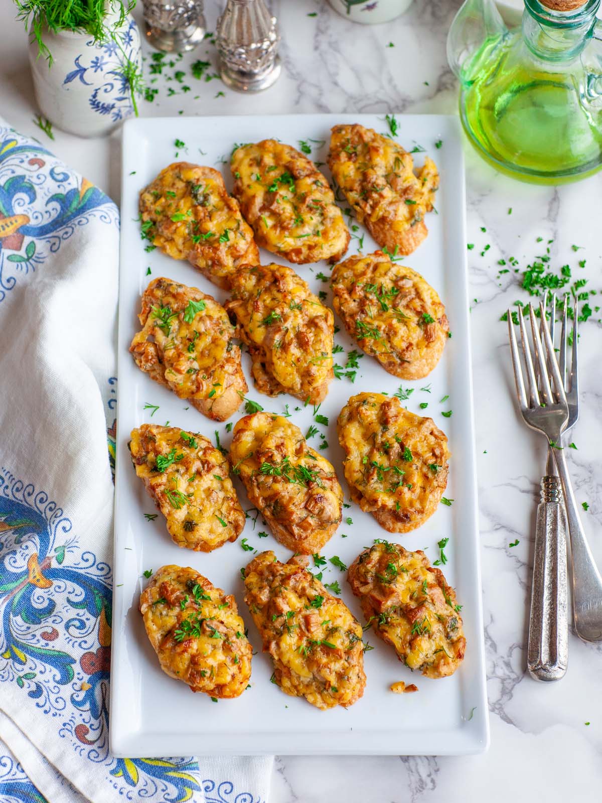 Many cheese mushroom crostini on a serving tray, on a table with fresh herbs and various kitchen items.