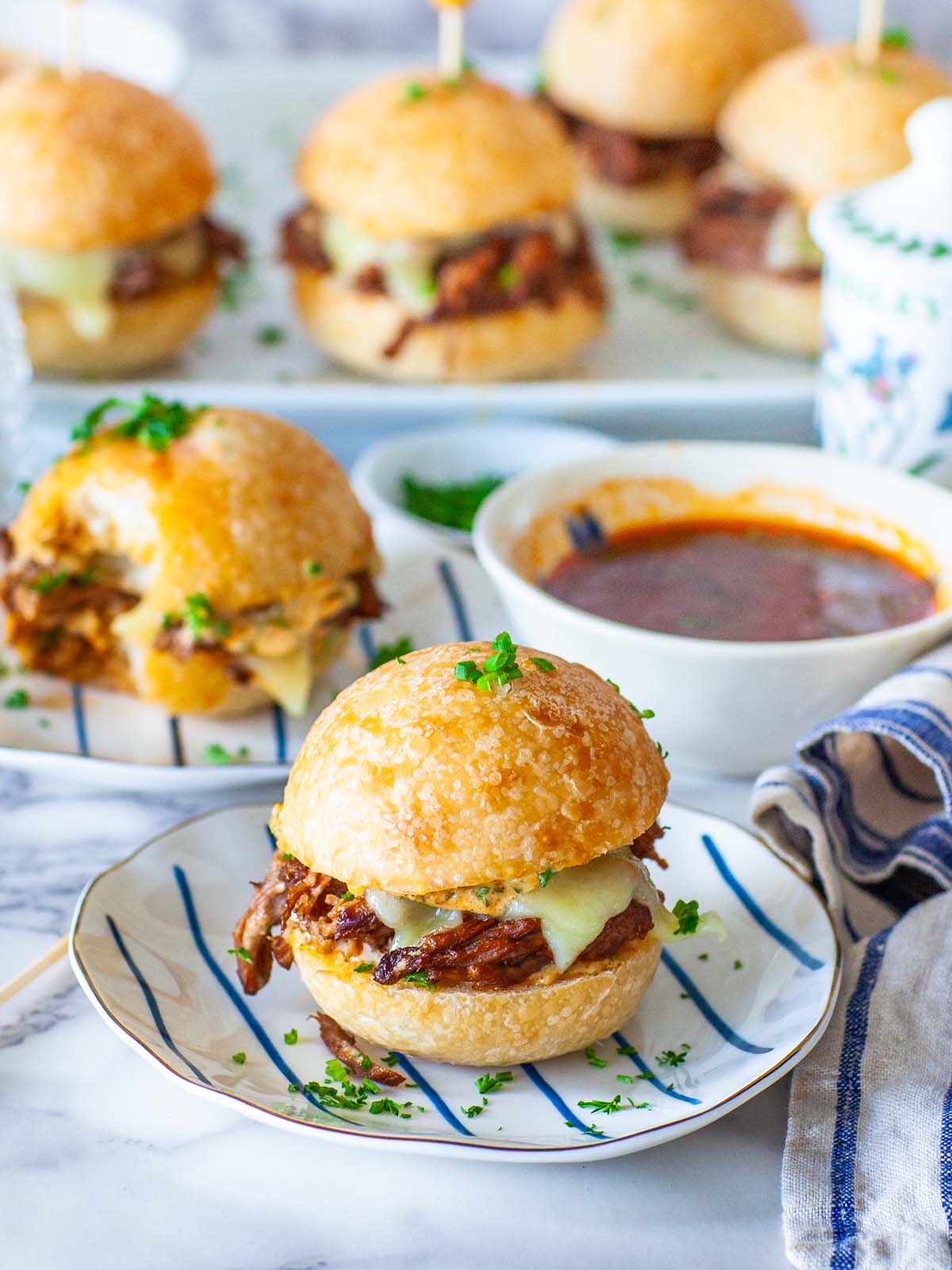 A beef slider sandwich on a plate, on a table with more sandwiches and dip.