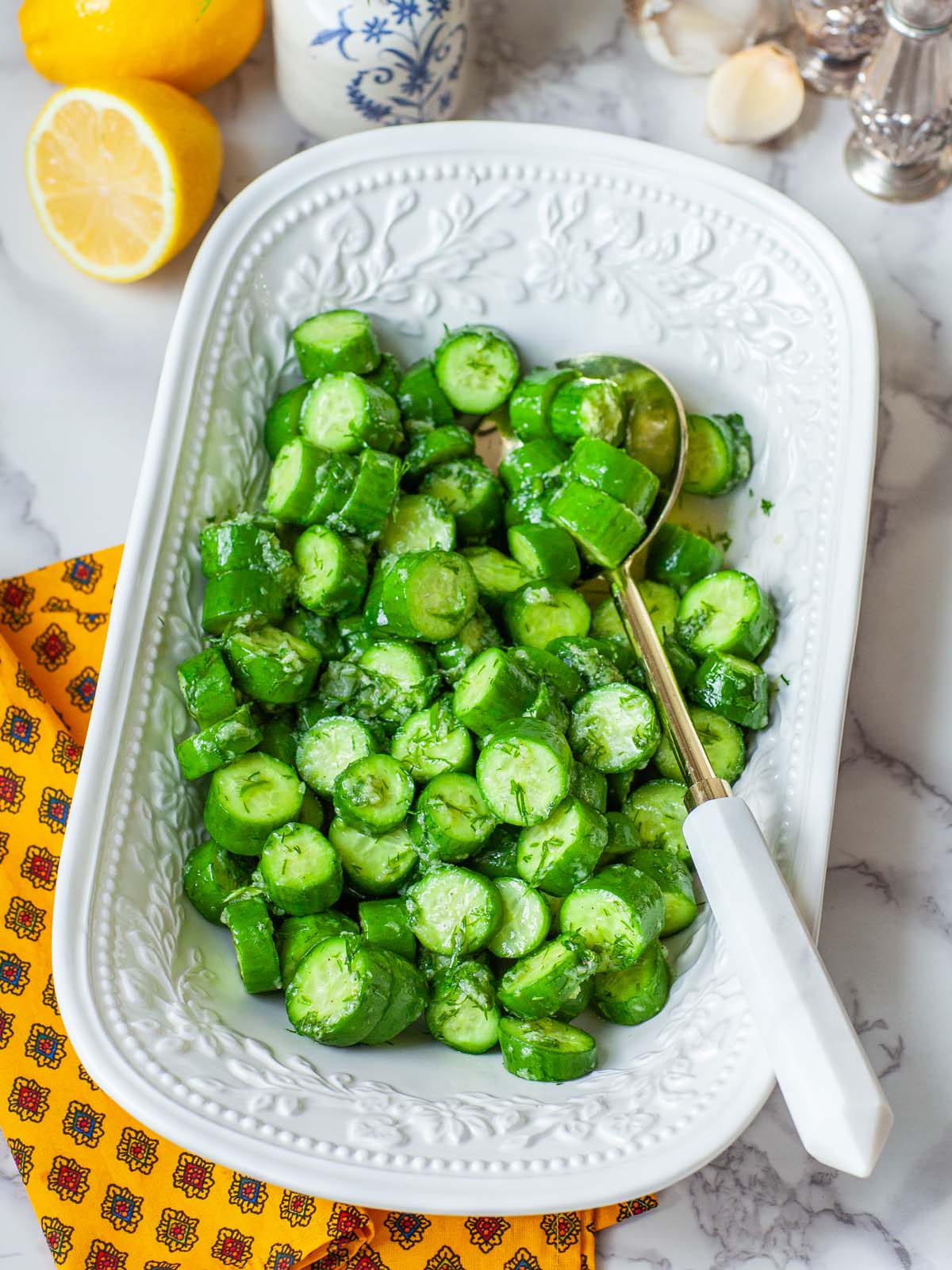 Chopped cucumbers in a serving bowl with a spoon, on a table with lemons and various kitchen items.