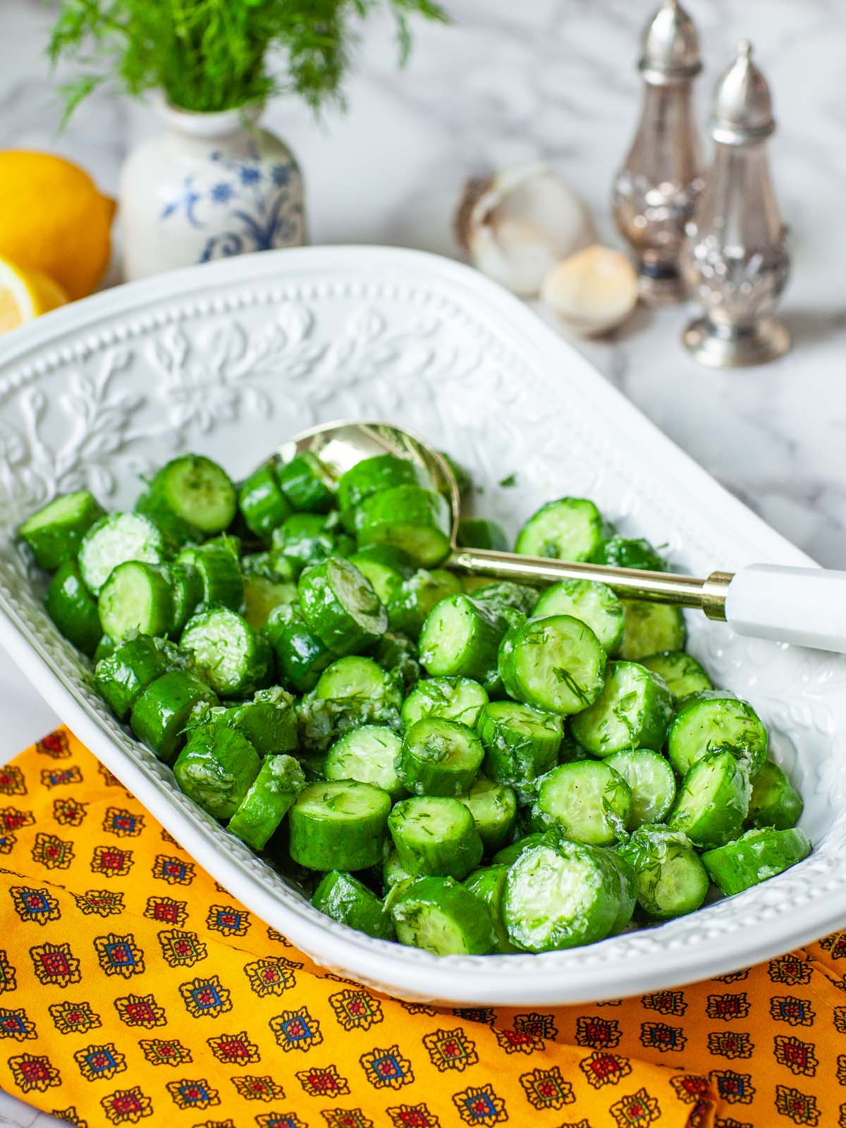 A closeup image of cucumber salad in a serving bowl, with utensils.