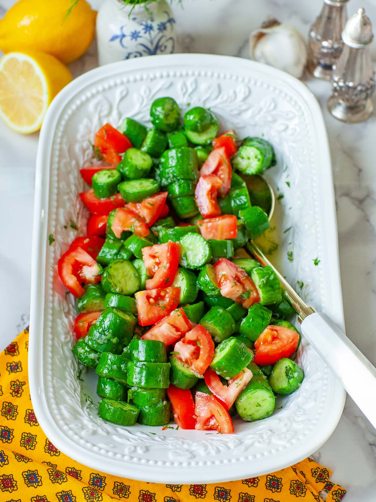 Tomato cucumber salad in a serving tray with a spoon, on a table with various kitchen items.