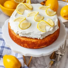 A simple lemon cake on a cake stand on a table with lemons and kitchen utensils.
