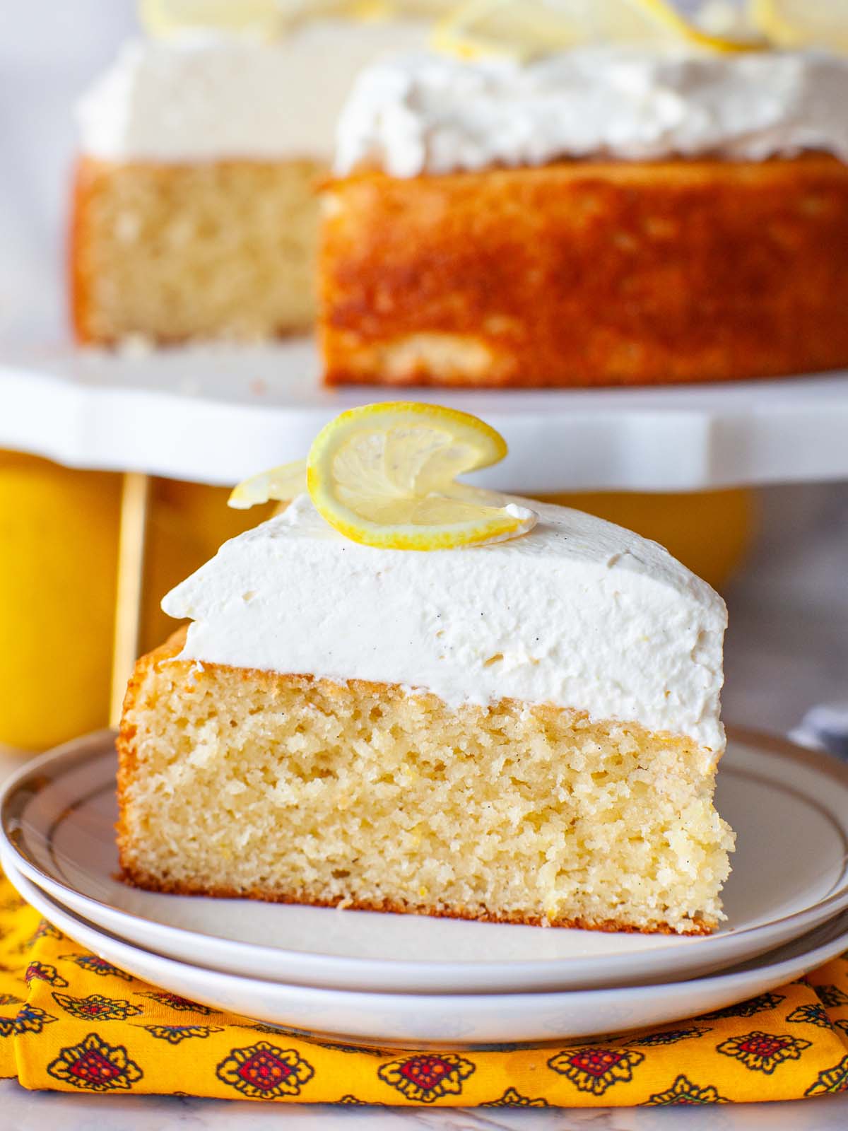 A closeup image of a cake slice with whipped cream and slice of lemon on top.