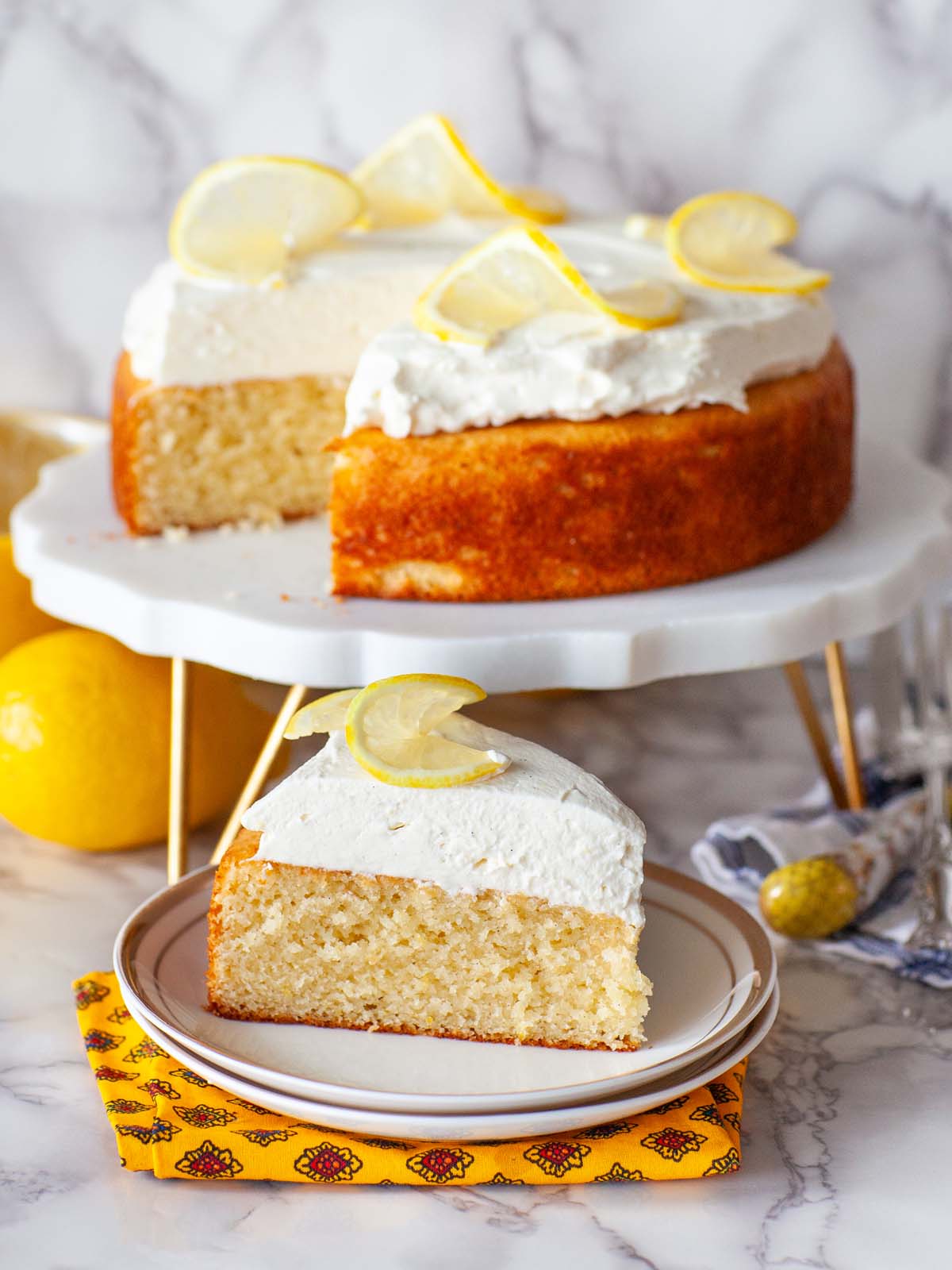 A closeup image of a slice of lemon cake on a plate, next to a cake on a cake stand.