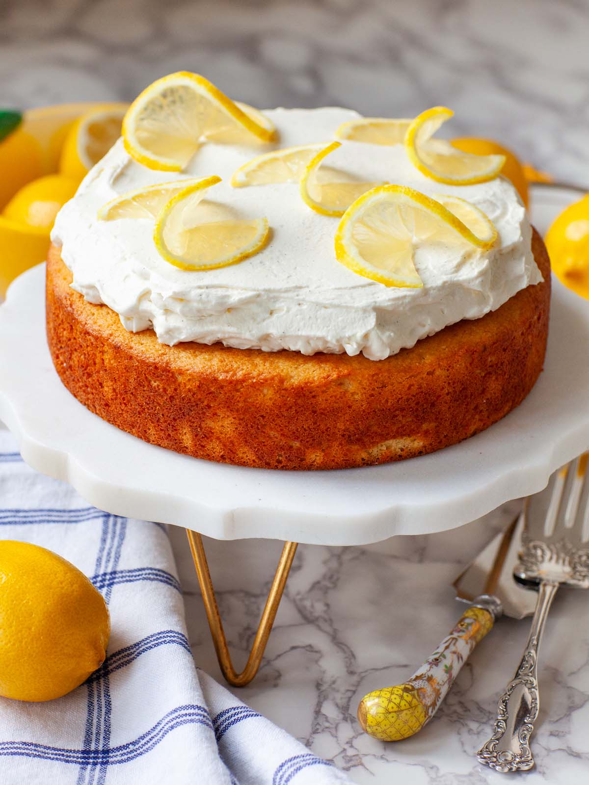 A cake topped with whipped cream and lemon slices on a cake stand, on a table with lemons and utensils.