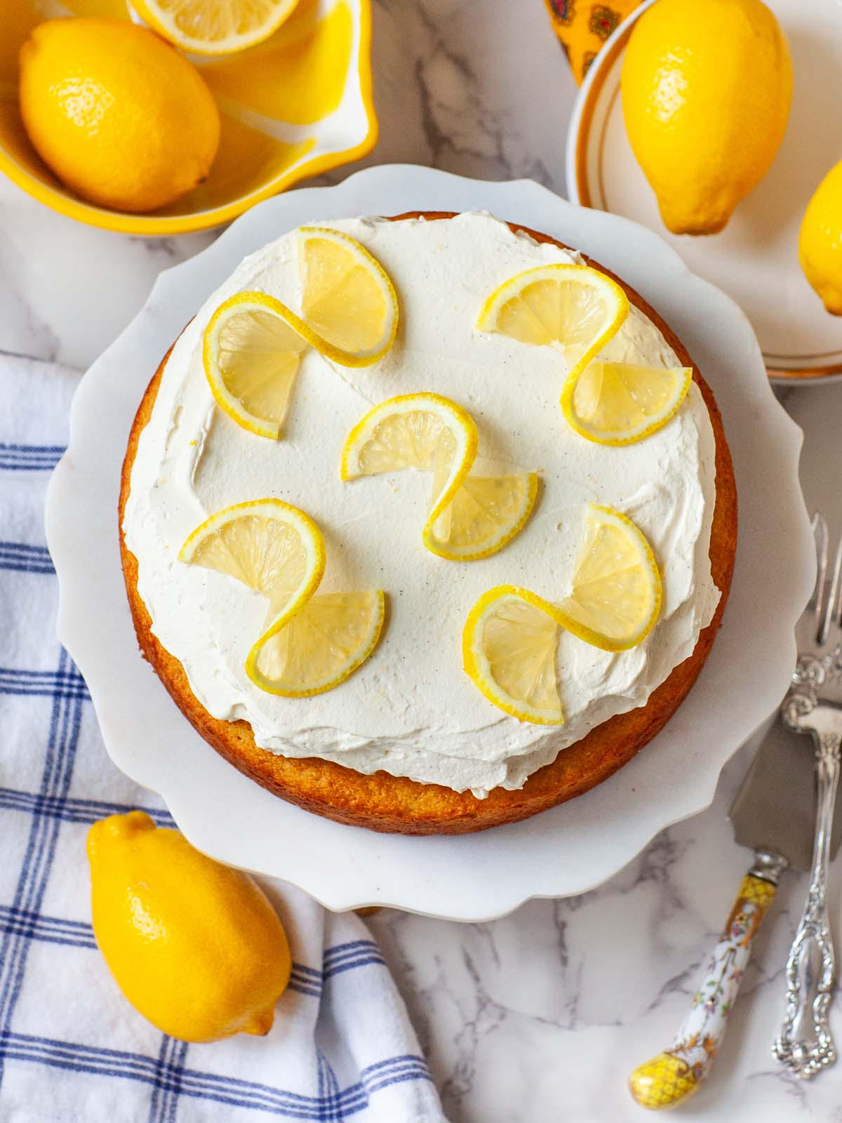 A lemon cake on a table with lemons, plates, and utensils.