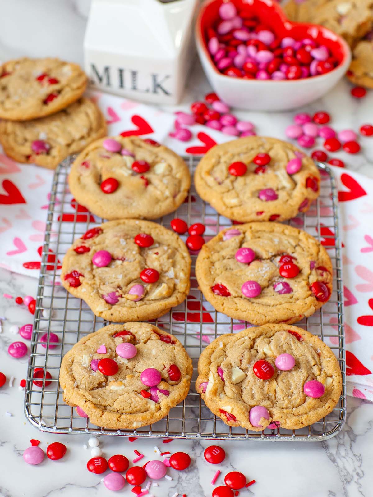 Brown cookies with pink and red M&Ms on a wire rack, on a table with more candy and cookies.