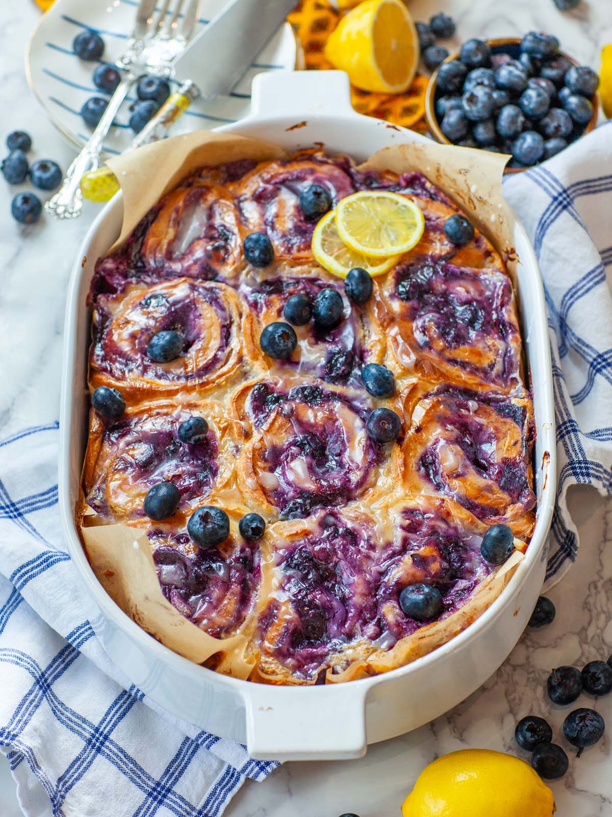 A casserole dish filled with blueberry rolls, on a table with lemons and blueberries.