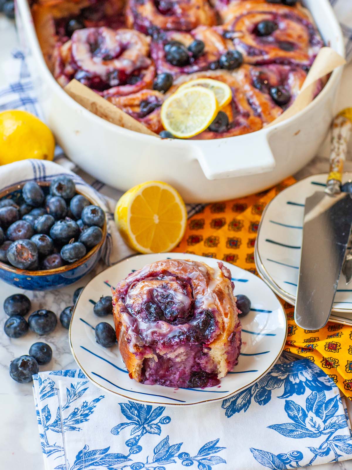 A blueberry cinnamon roll on a plate, on a table with a casserole dish, berries, and lemons.