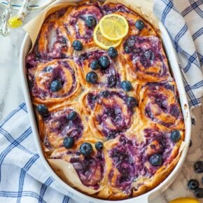 A casserole dish filled with blueberry rolls, on a table with lemons and berries.
