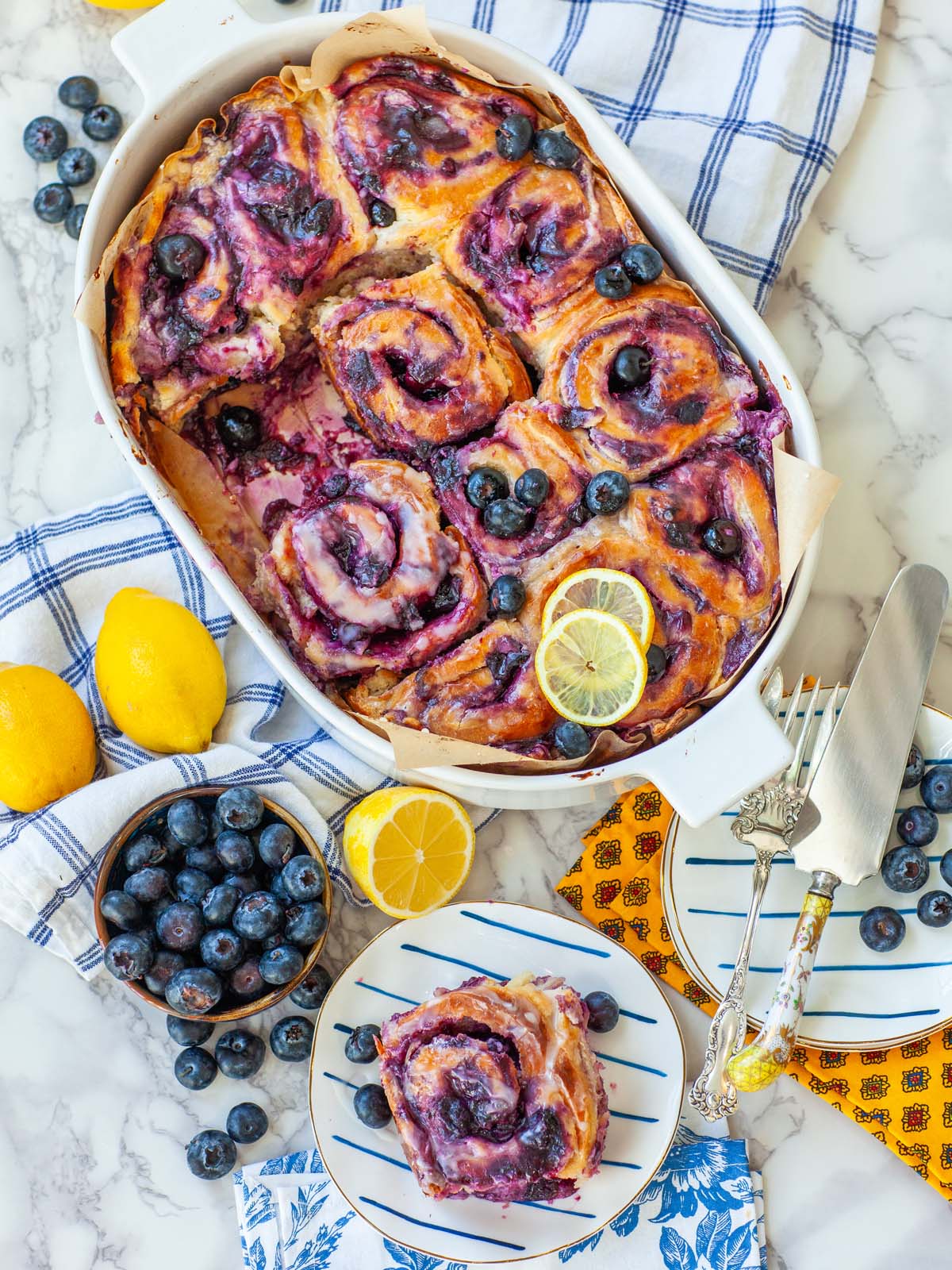 A casserole filled with blueberry cinnamon rolls on a table with lemons, blueberries, and a roll on a plate.
