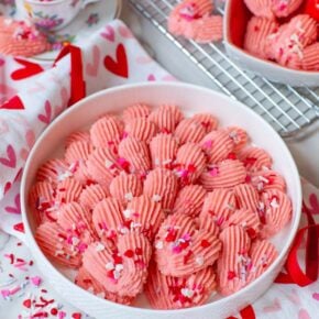 A plate filled with heart-shaped pink cookies with sprinkles, on a table with more cookies and kitchen items.