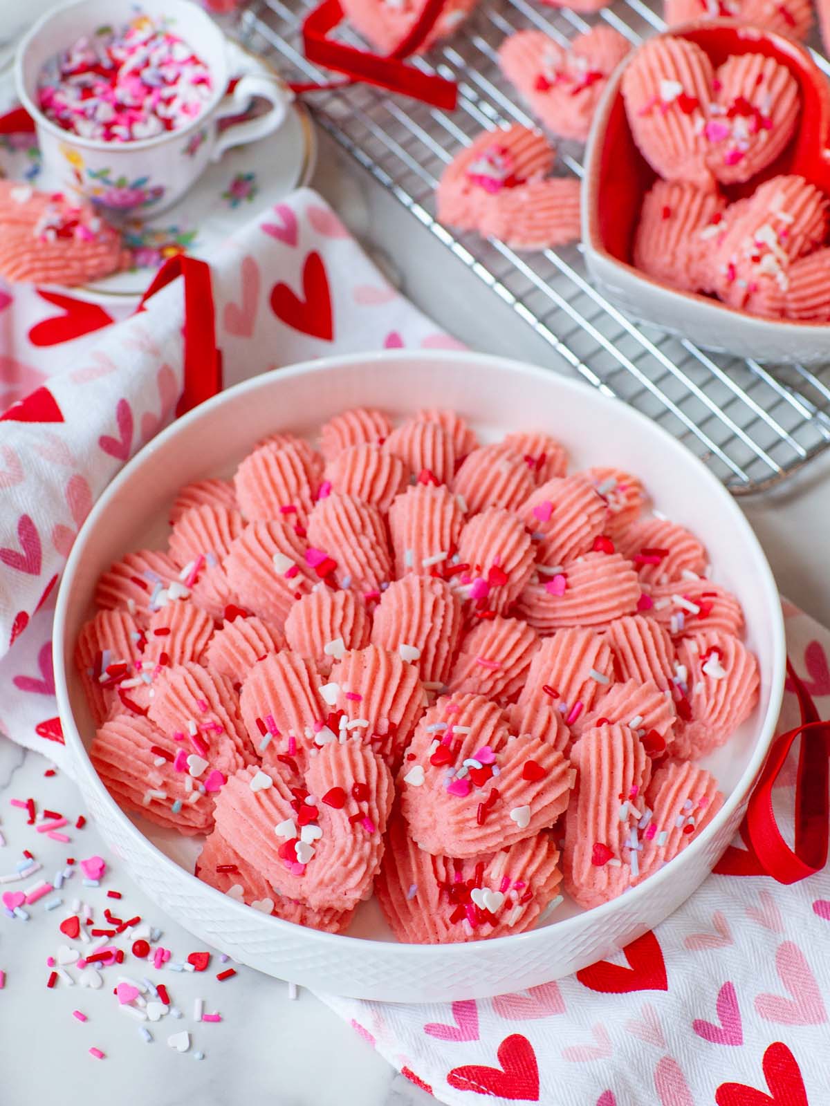 A plate filled with heart-shaped pink cookies with sprinkles, on a table with more cookies and kitchen items.