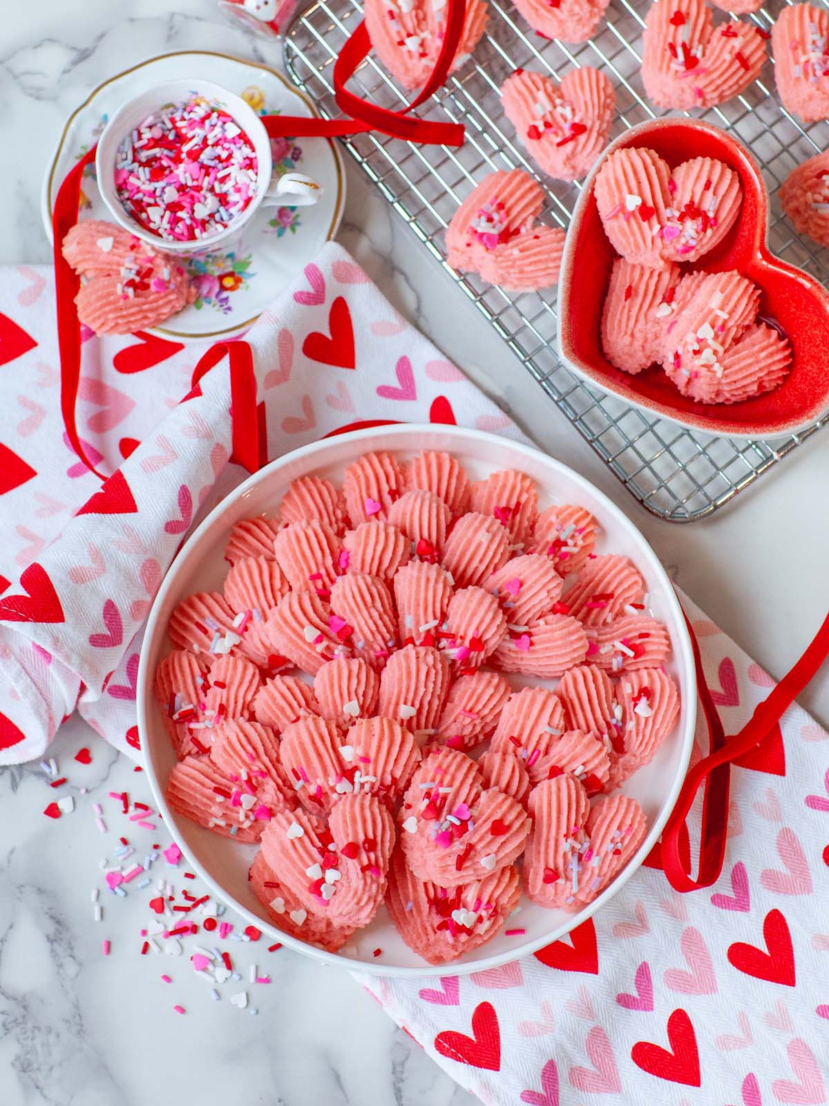 A tray of pink, heart-shaped cookies on a table with a wire cooling rack, more cookies, tea cup, and tea towel with hearts.