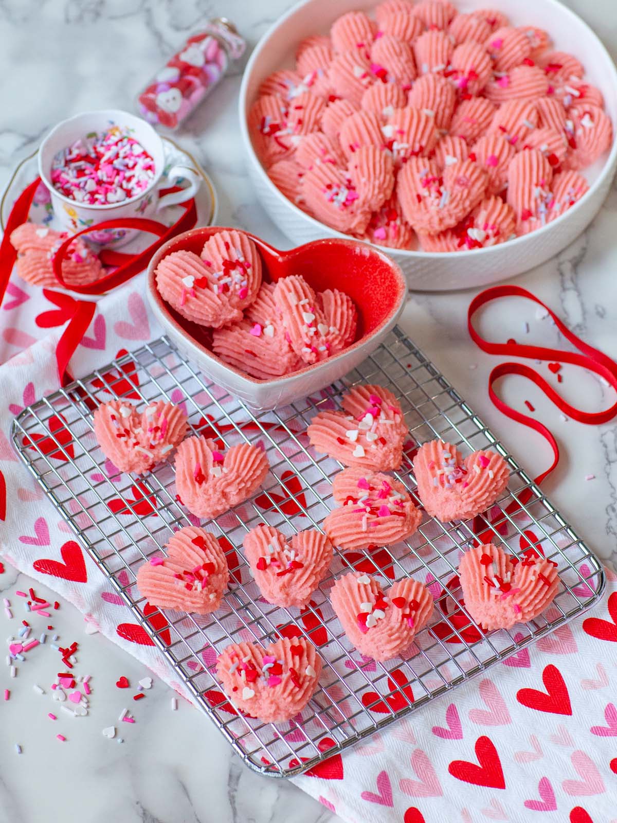 Pink, heart-shaped cookies on a cooling rack, on a table with a tray of cookies.