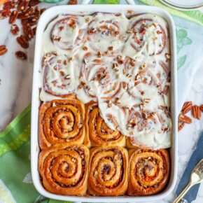 A casserole dish filled with carrot cake cinnamon rolls with icing, on a table with various bowls and utensils.
