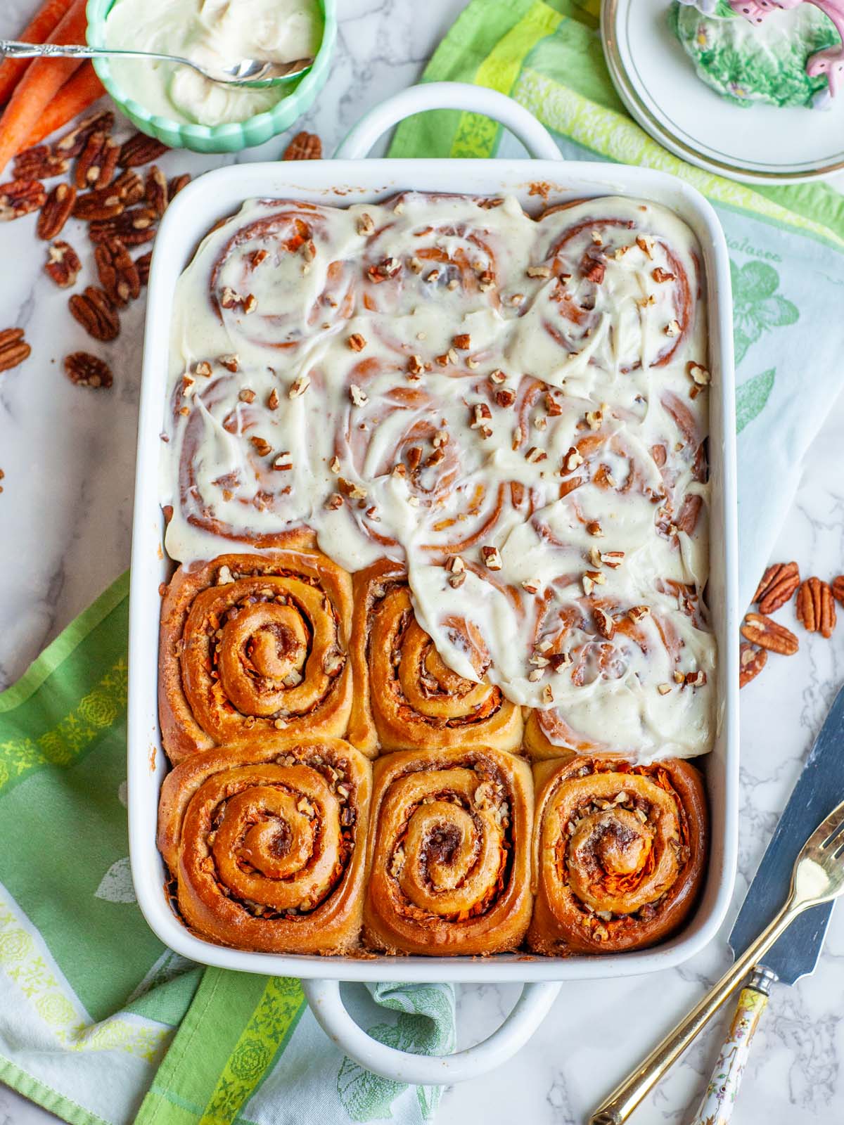 A casserole dish filled with carrot cake cinnamon rolls with icing, on a table with various bowls and utensils.