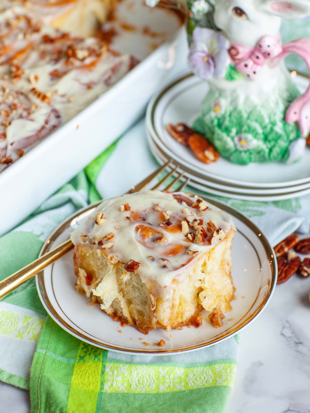 A closeup image of a cinnamon roll on a plate, on a table with various items.