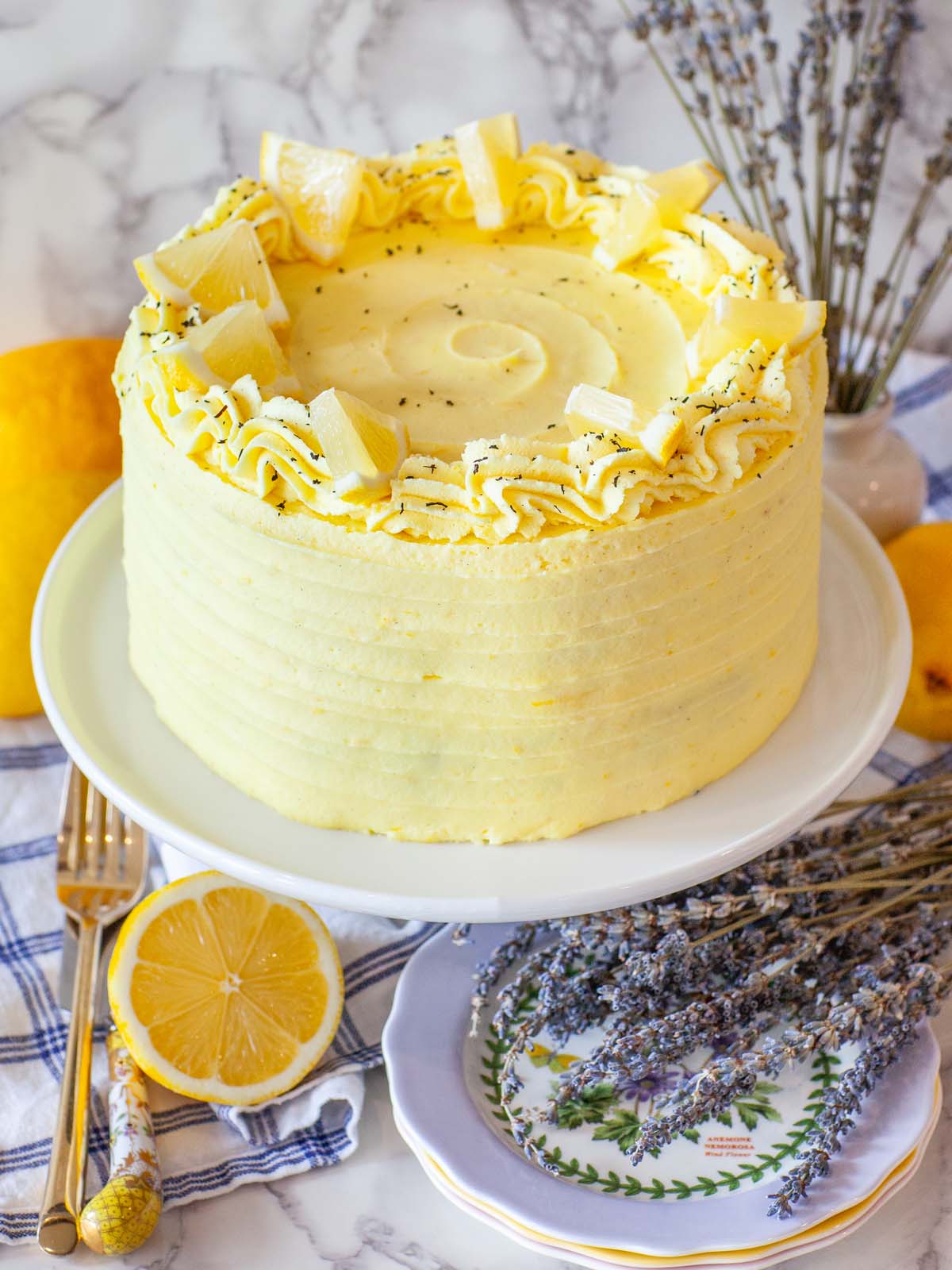 A lemon yellow cake on a cake stand, with lemons and lavender flowers on the table below.