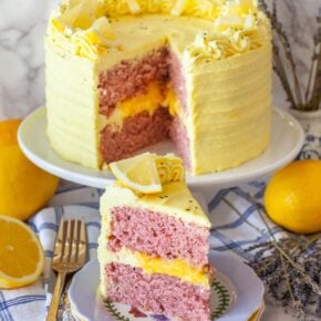 A slice of cake on a plate next to a cake on a cake stand, on a table with lemons and lavender.