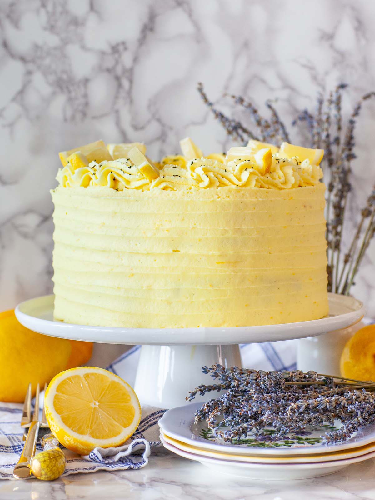 A yellow lemon cake on a cake stand, on a table with lemons, lavender flowers, and plates.