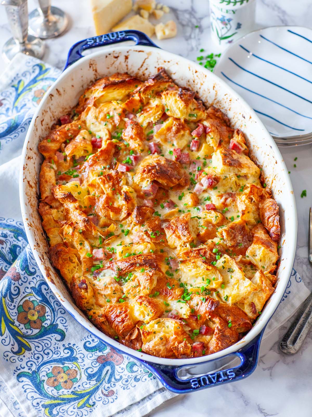 An oval casserole dish with croissant casserole, dotted with ham; on a table with plates and utensils.