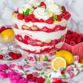 A glass trifle bowl filled with cream and raspberries, topped with berries. On a table with raspberries and lemons.