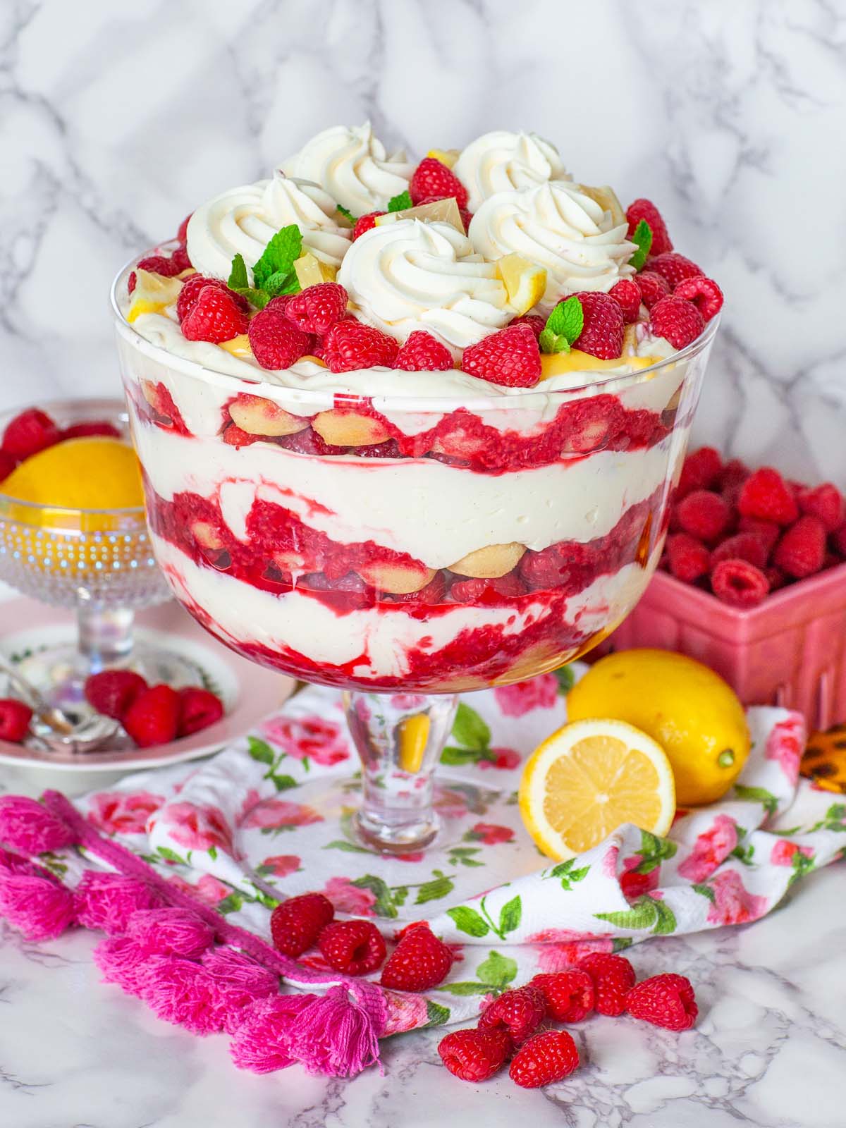 A glass trifle bowl filled with cream and raspberries, topped with berries. On a table with raspberries and lemons.