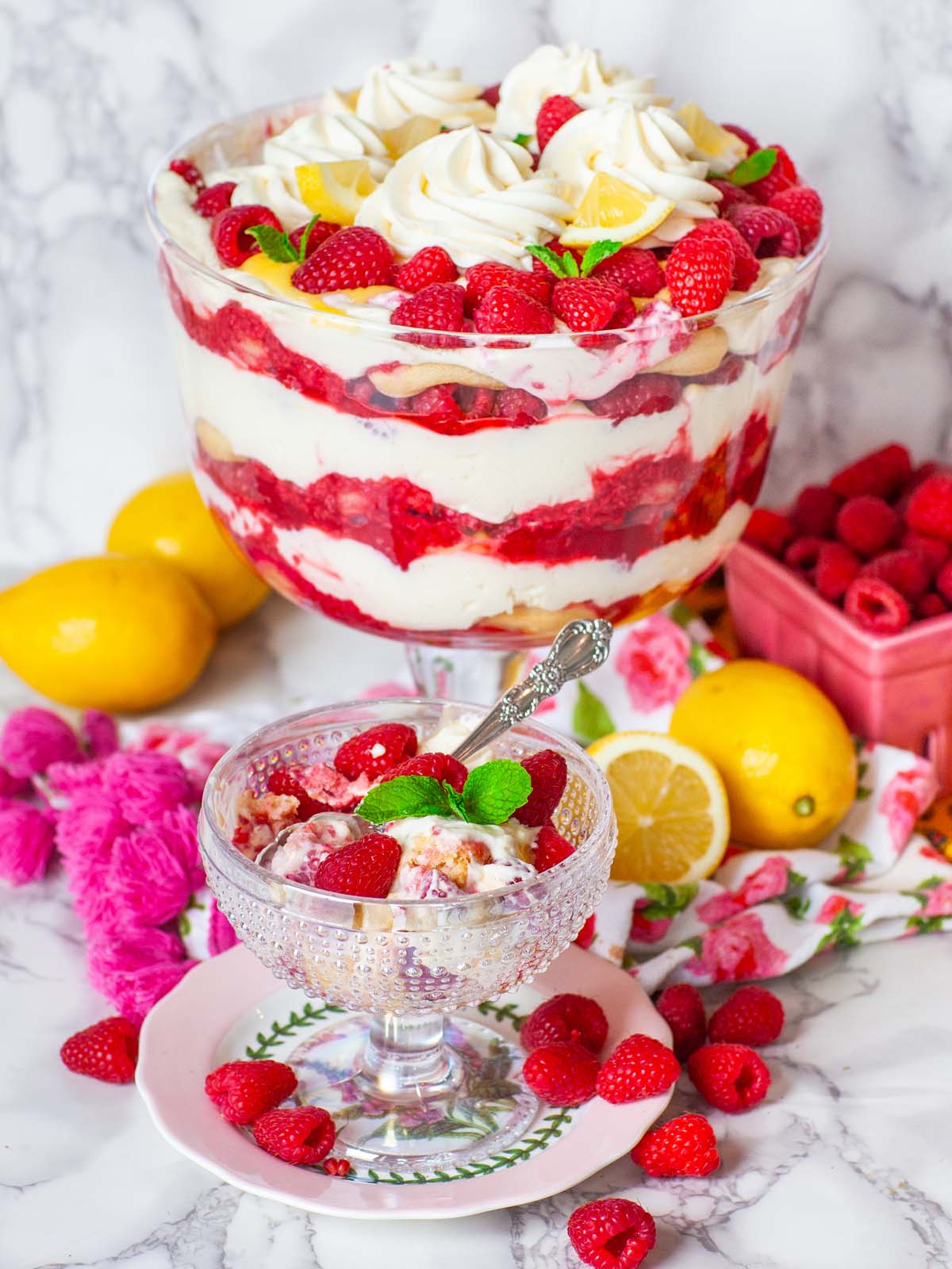 A small bowl filled with cream and raspberries, on a table with a large raspberry trifle bowl. On a table with lemons and raspberries.