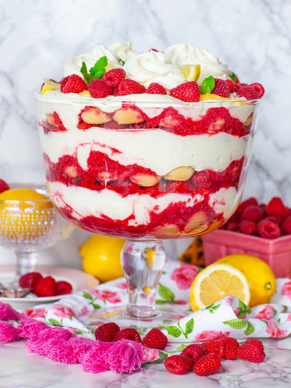 A glass trifle bowl filled with layers of cream, cookies, and raspberries. On a table with raspberries and lemons.